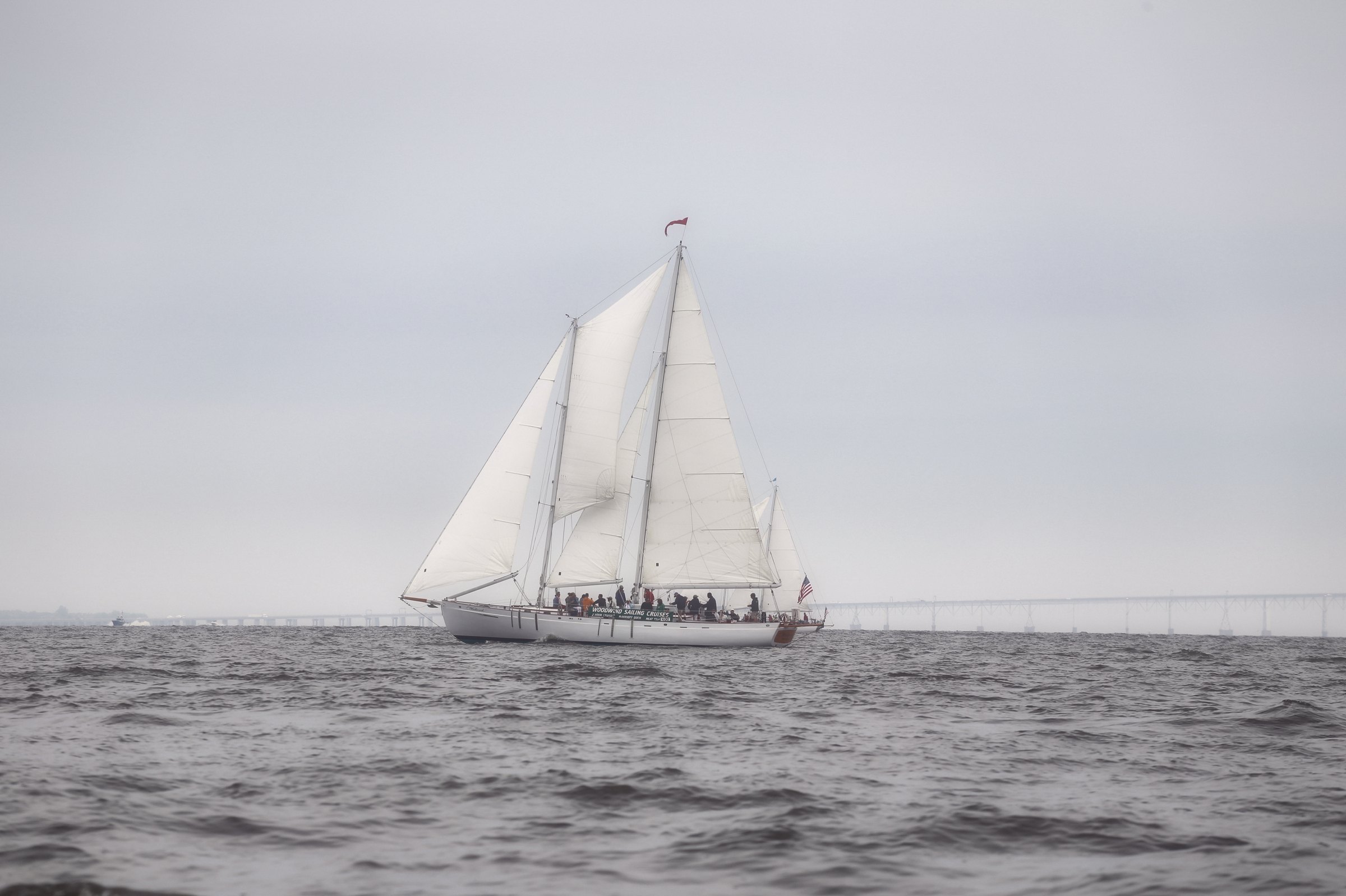 A two-masted schooner under full sail on a soft grey bay.