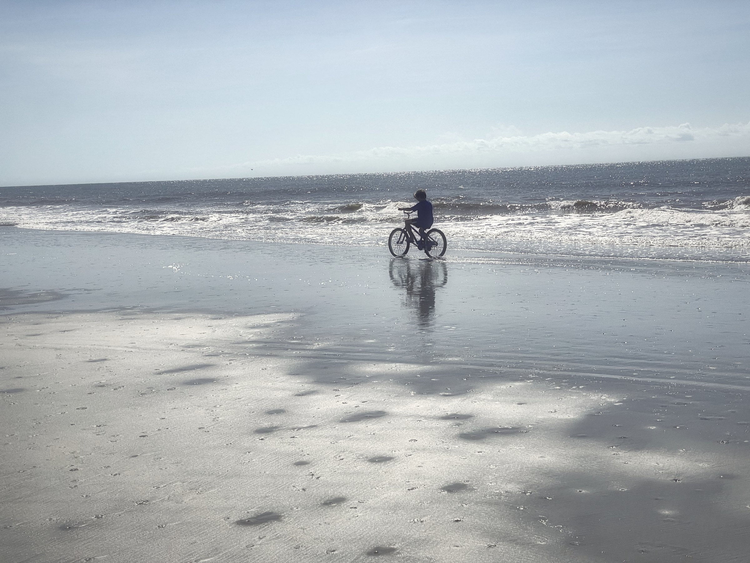 A lone cyclist on wet, reflective sand at the edge of the surf.
