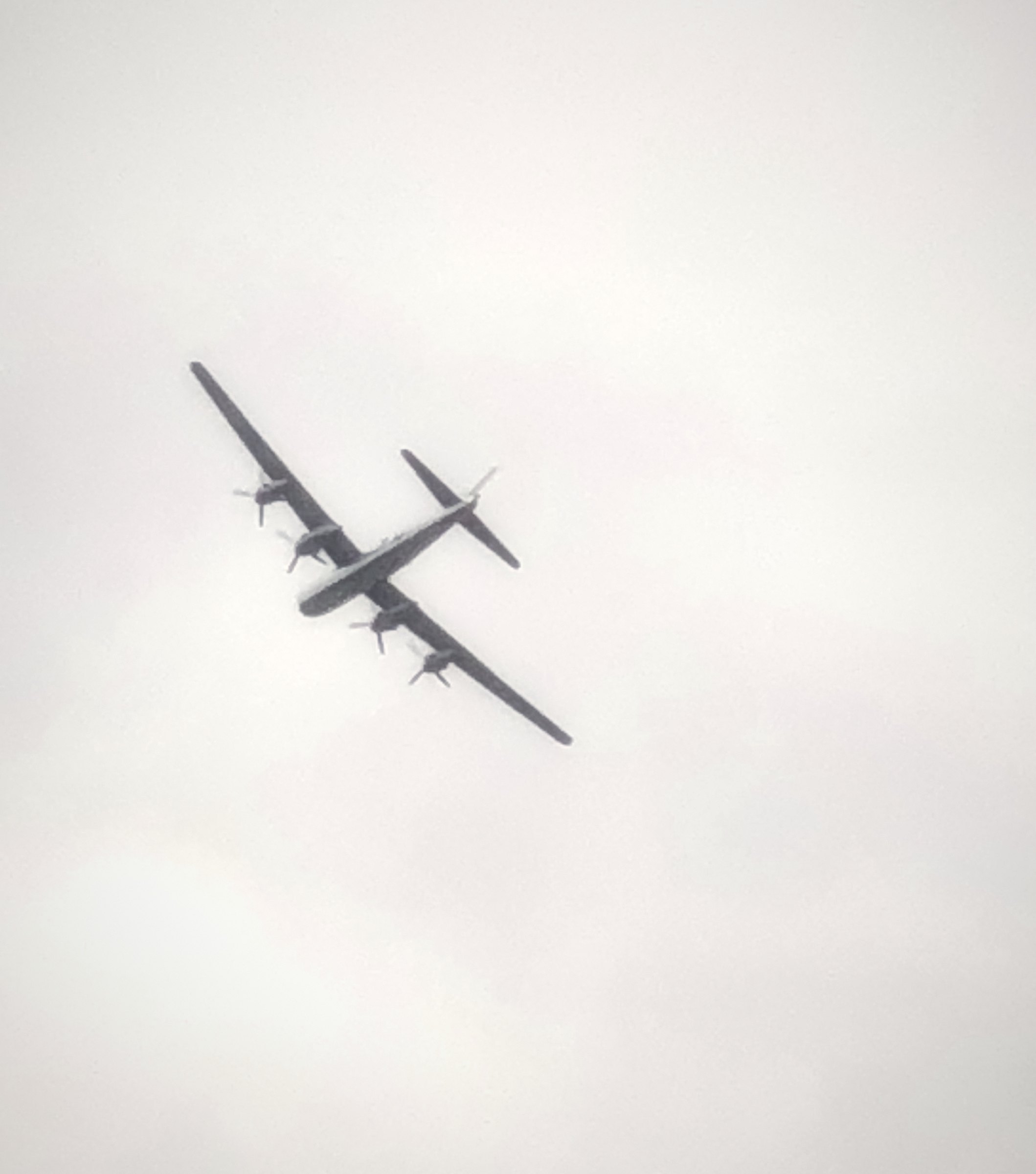 A four-engine warbird banking through a white, hazy sky.