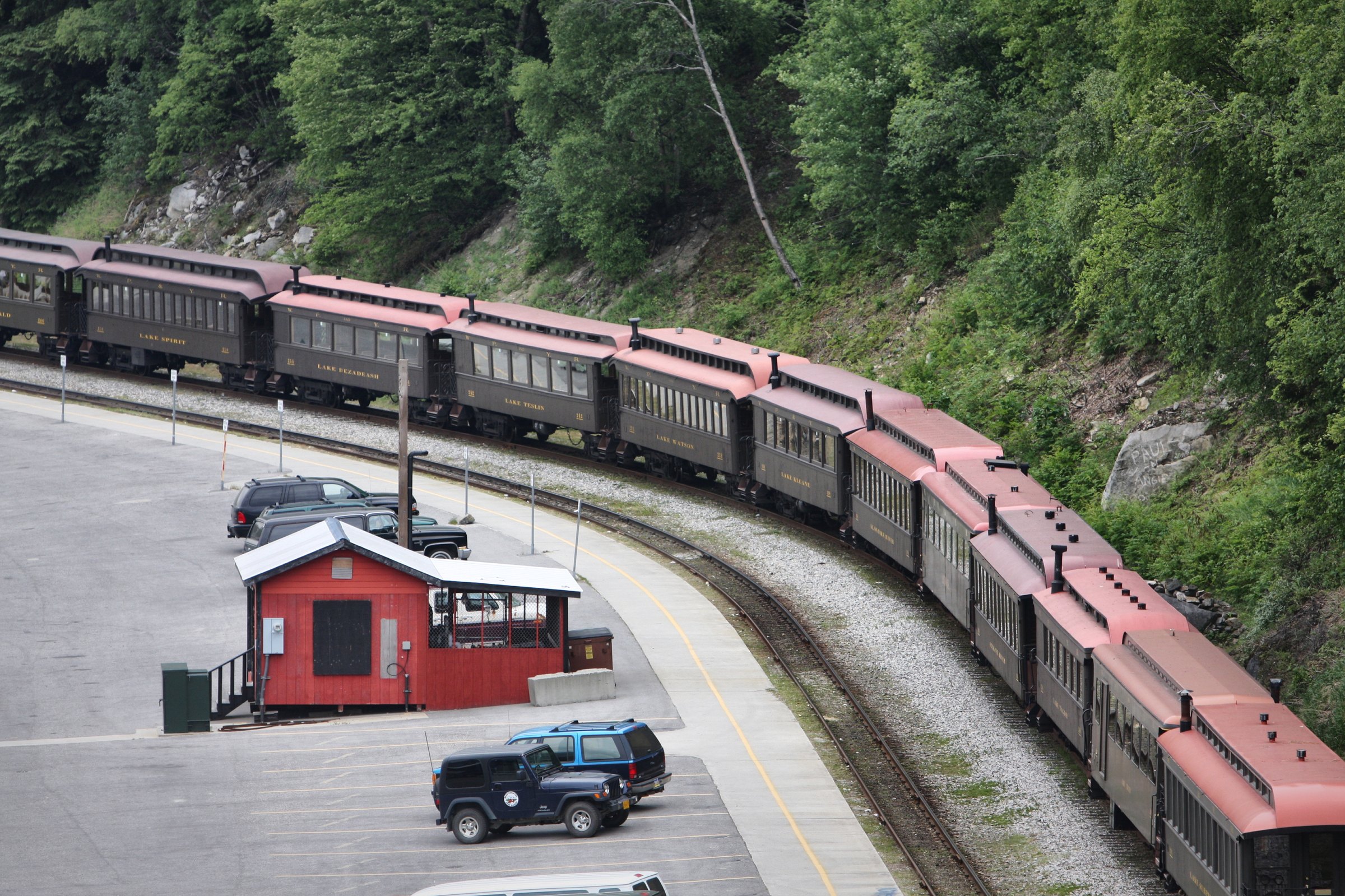 A long line of vintage passenger cars with rose-red roofs curving along a mountainside track.