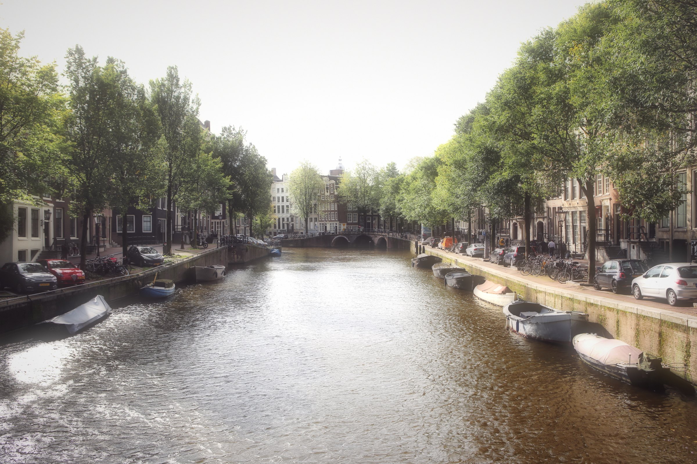 Moored boats along a tree-shaded canal under a hazy sky.