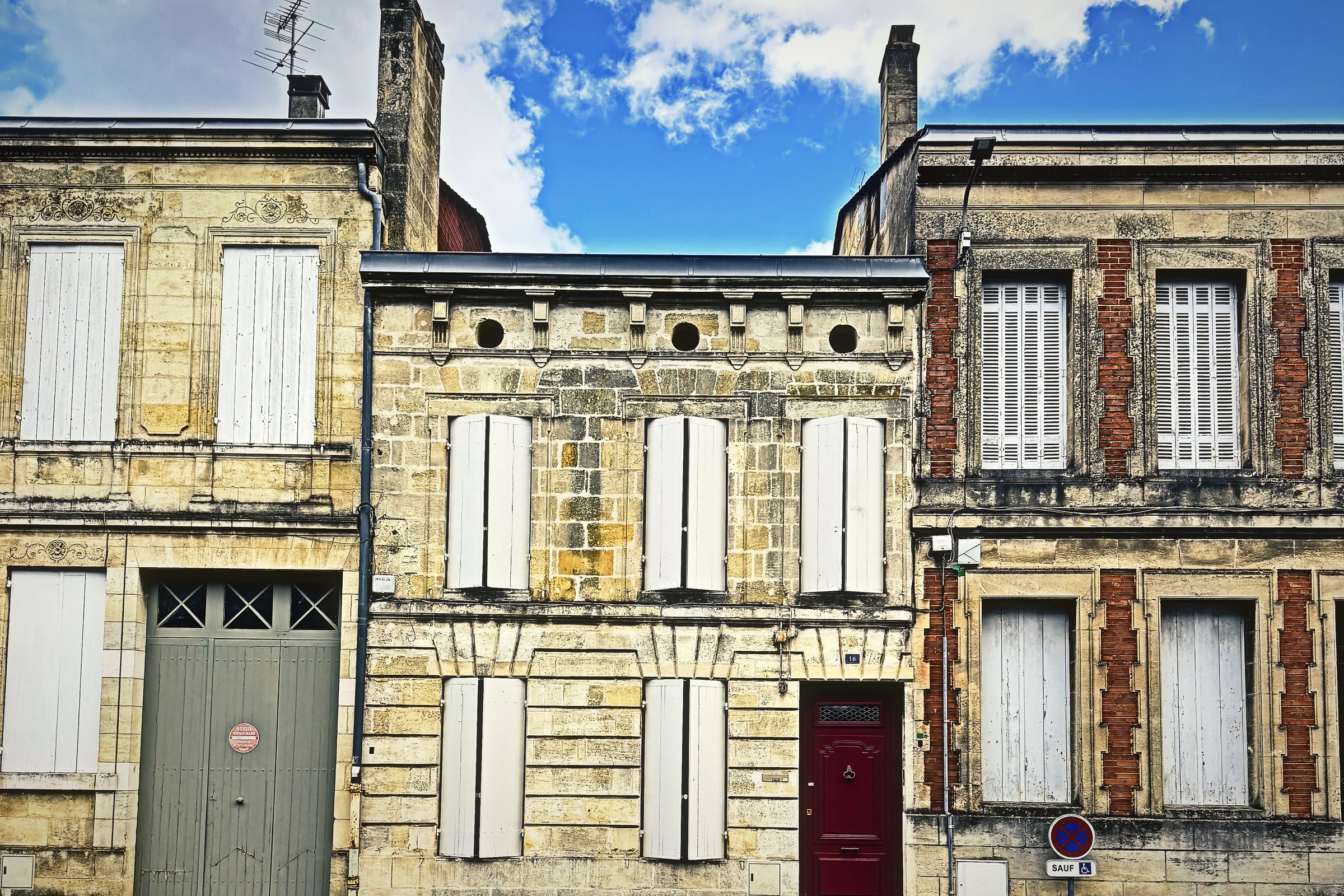 Three stone townhouse fronts side by side with pale shutters, a grey garage door and a red door.