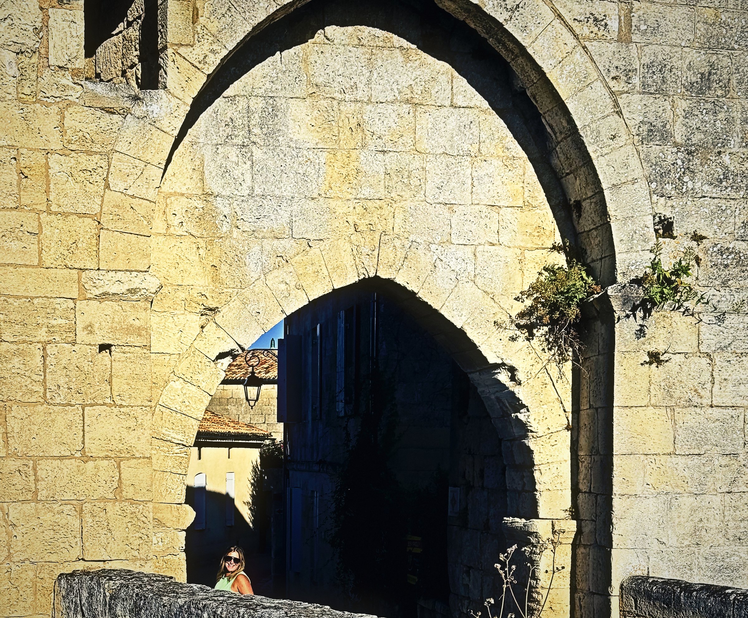 A tall pointed stone arch in sunlit limestone above a shadowed passage and a low wall.