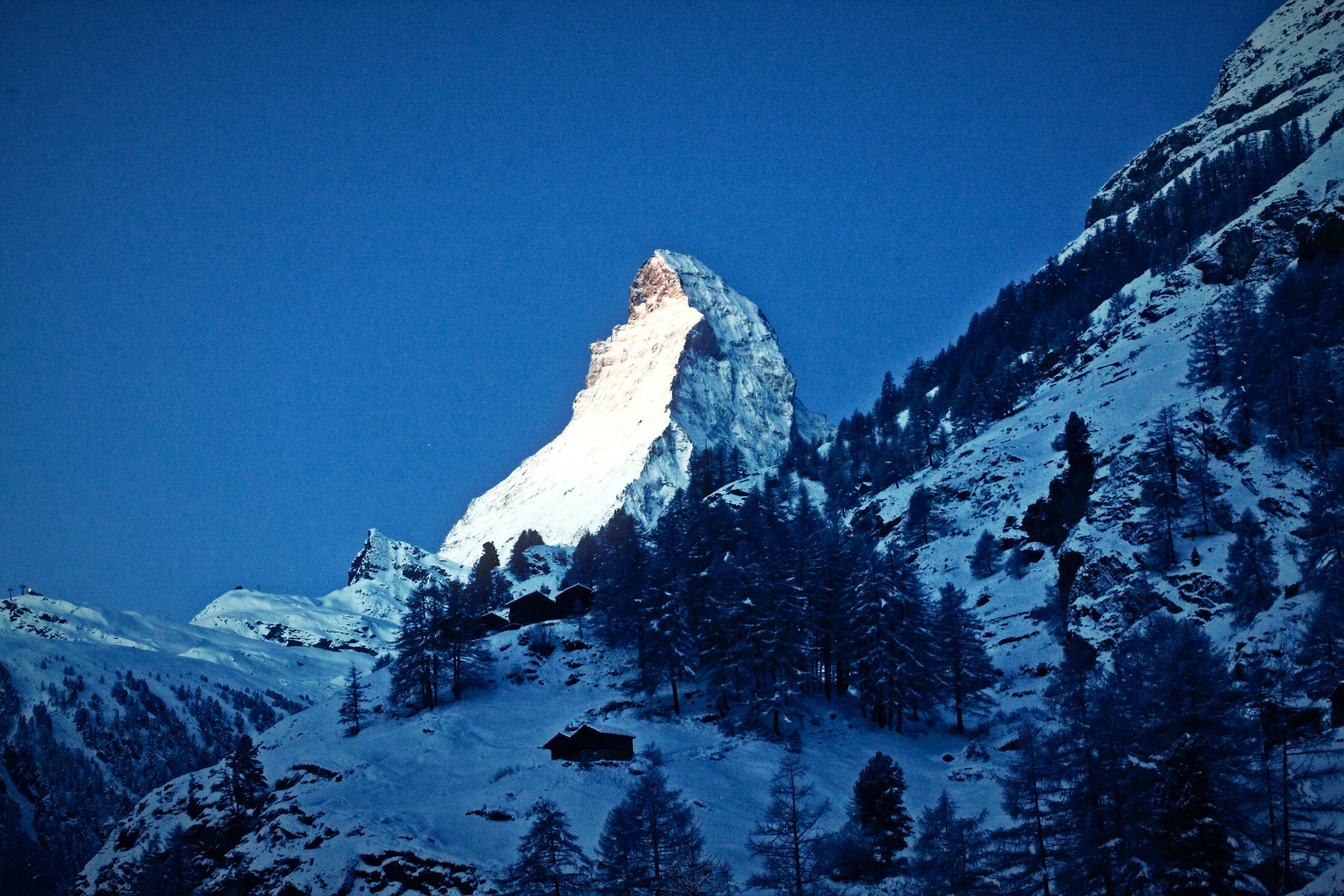 Matterhorn summit lit pink and gold at dawn above a snowy forest and a deep blue valley.
