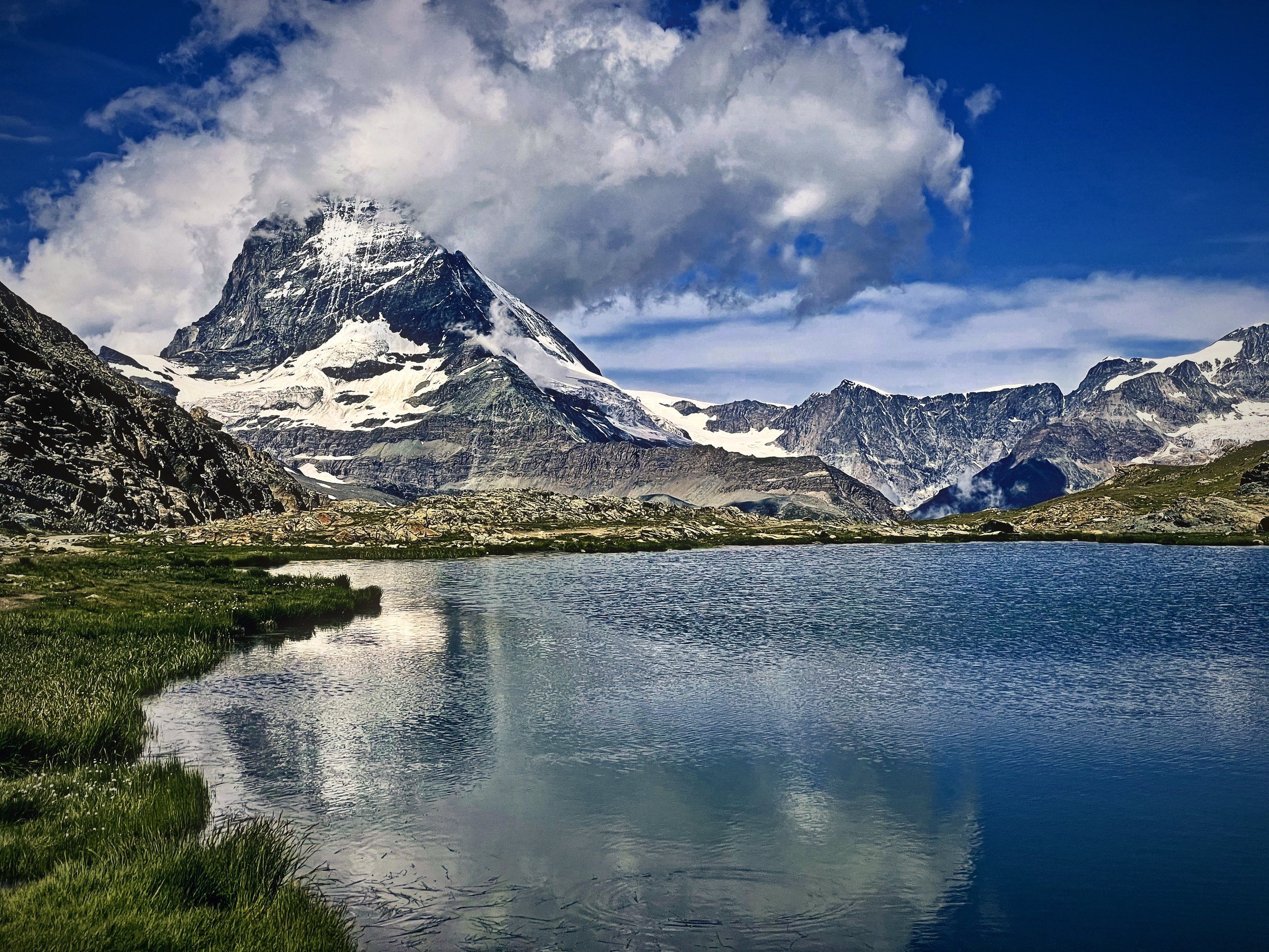 The Matterhorn and its clouds reflected in a still alpine lake edged with green meadow.