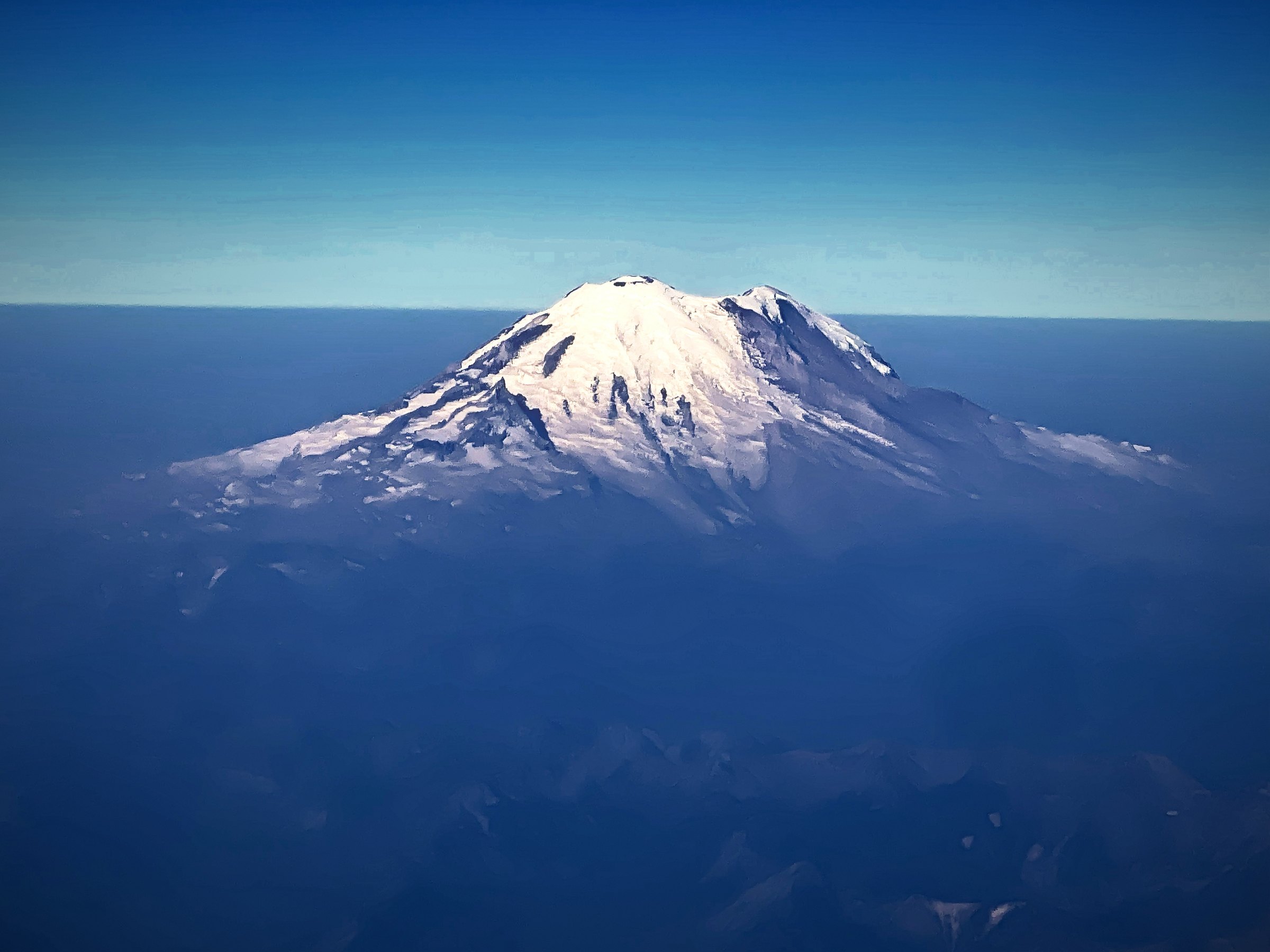 Snow-capped volcano rising alone above a sea of blue haze, seen from the air.