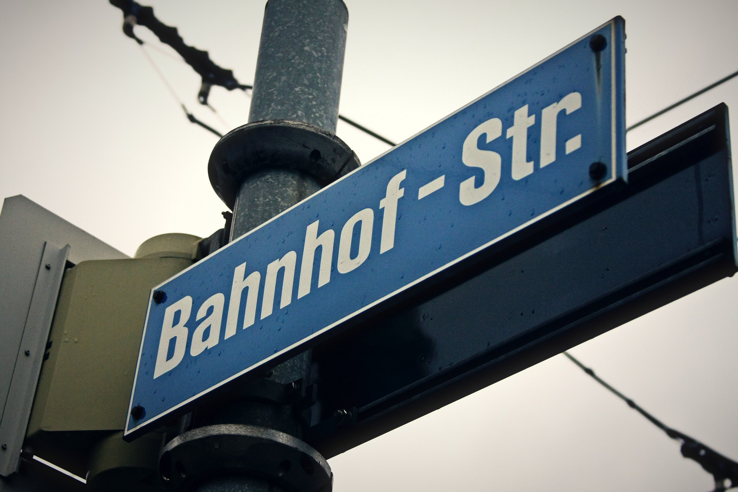 Blue street sign reading Bahnhof-Str. on a post under tram wires.