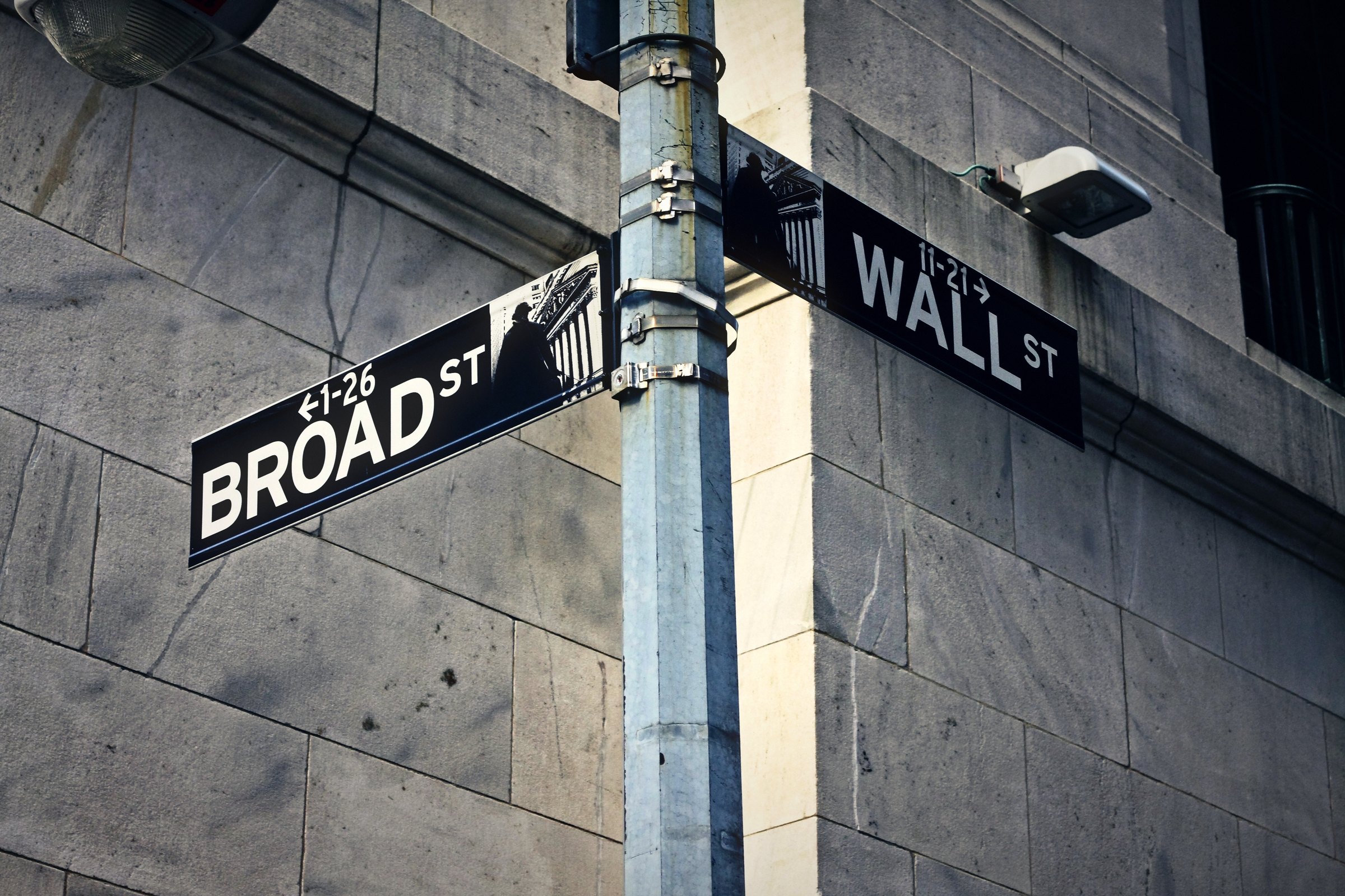 Two black street signs reading Broad St and Wall St on a lamppost against stone.