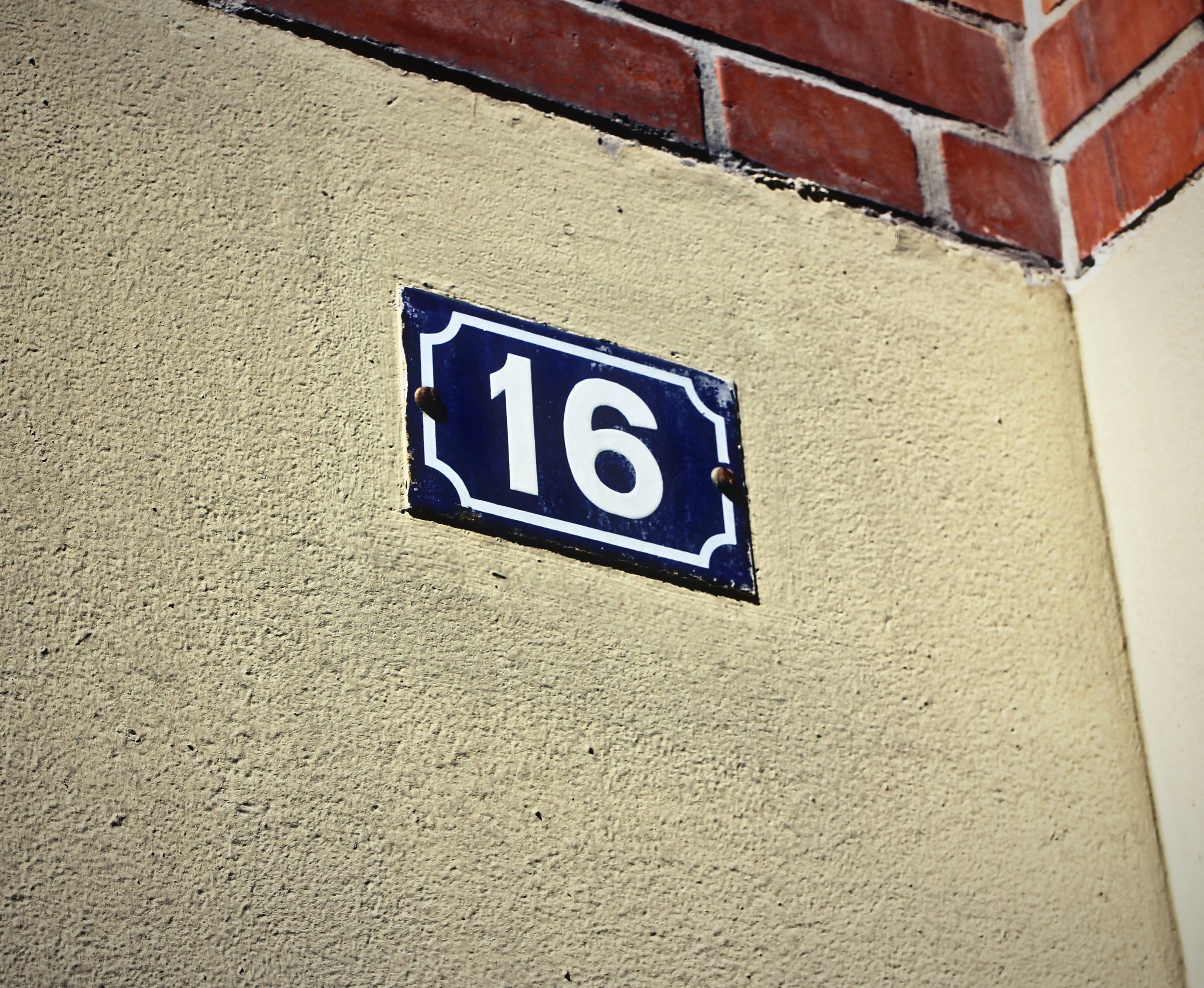 Small blue enamel house number 16 on a cream wall under brick.