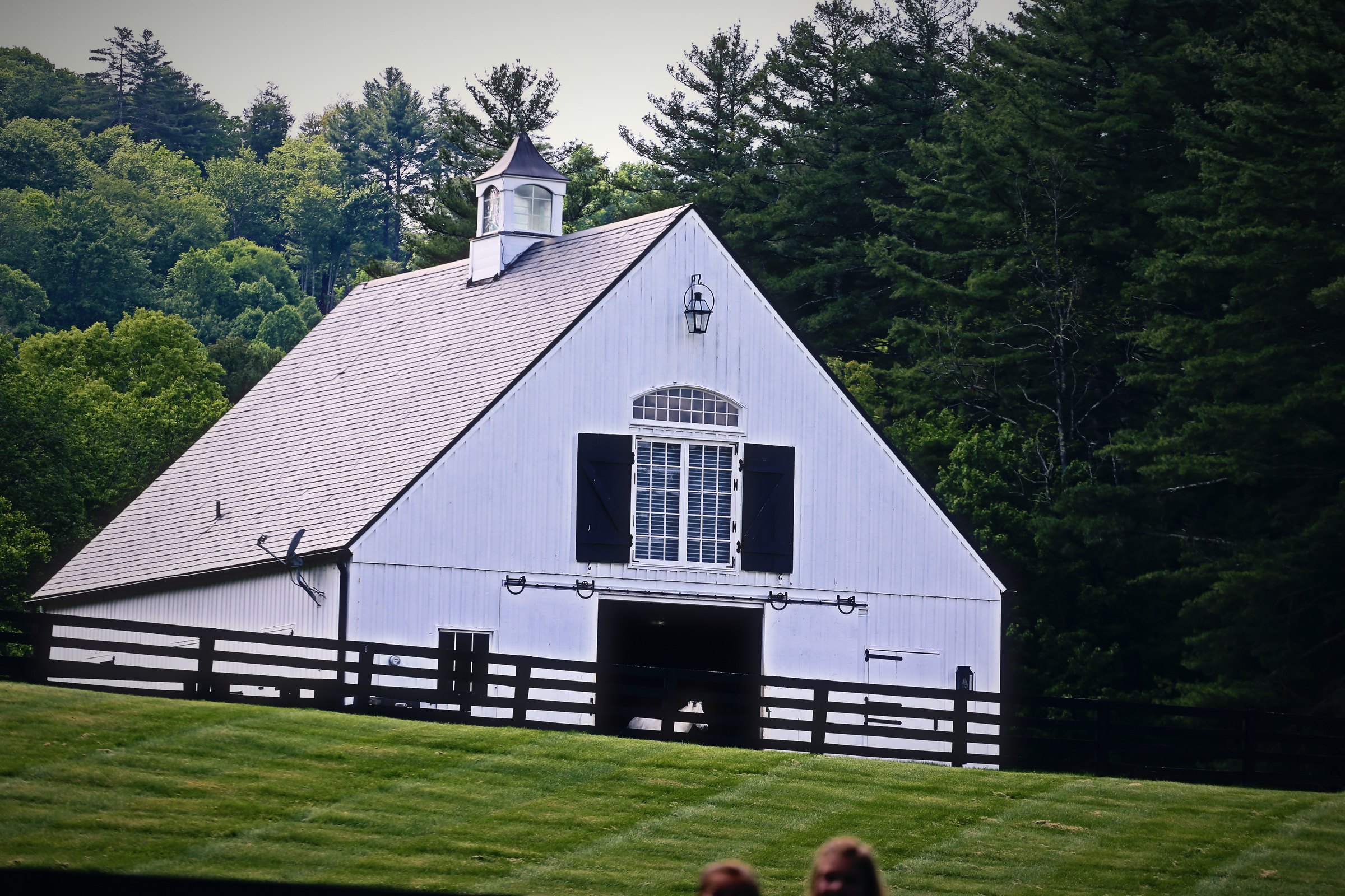 A white barn with a steep roof and cupola framed by dark green forest.