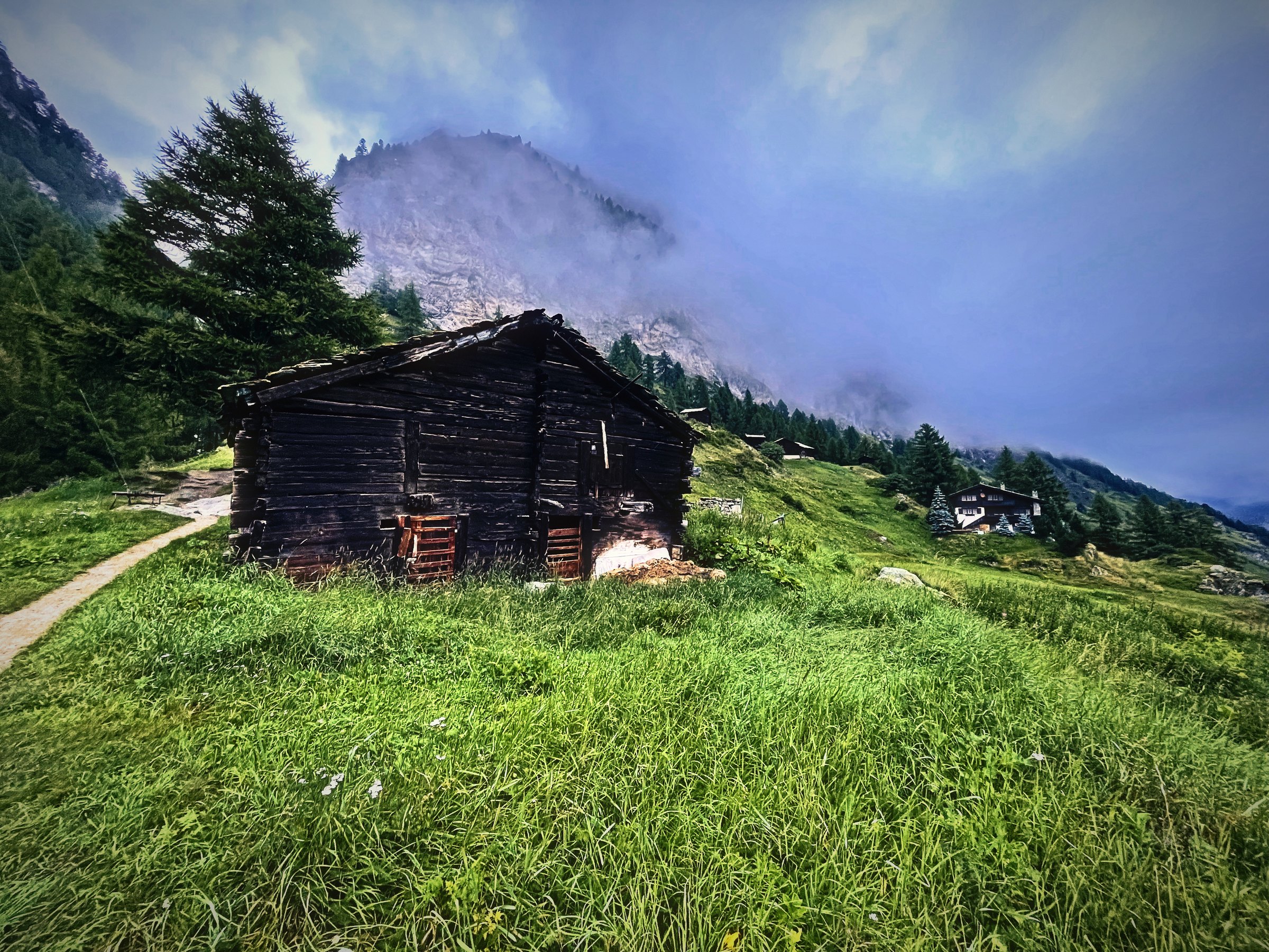 A dark weathered wooden hut on a green alpine slope beneath cloud-wrapped peaks.