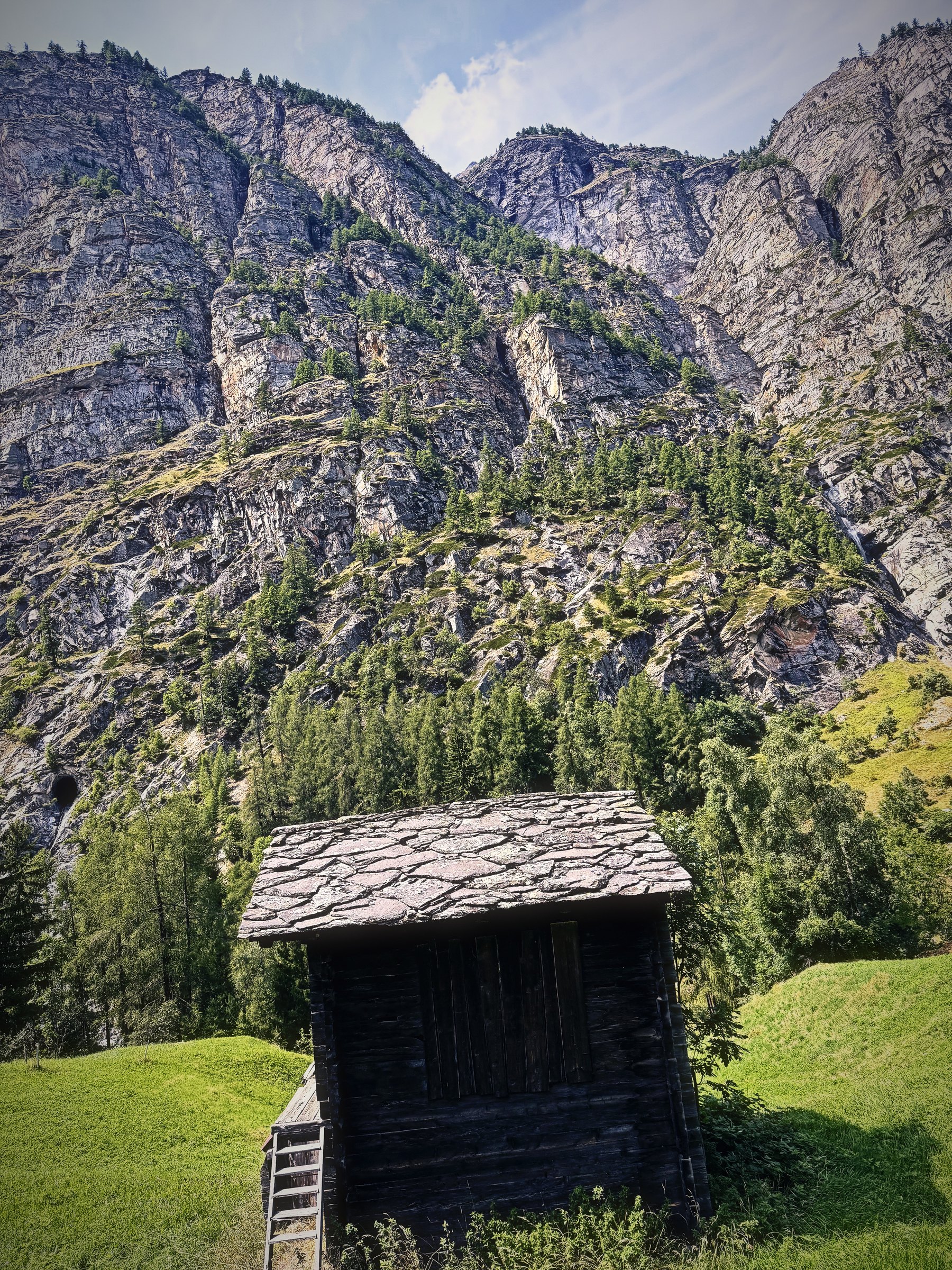 A small dark wooden storage hut with a stone roof below towering rock walls.