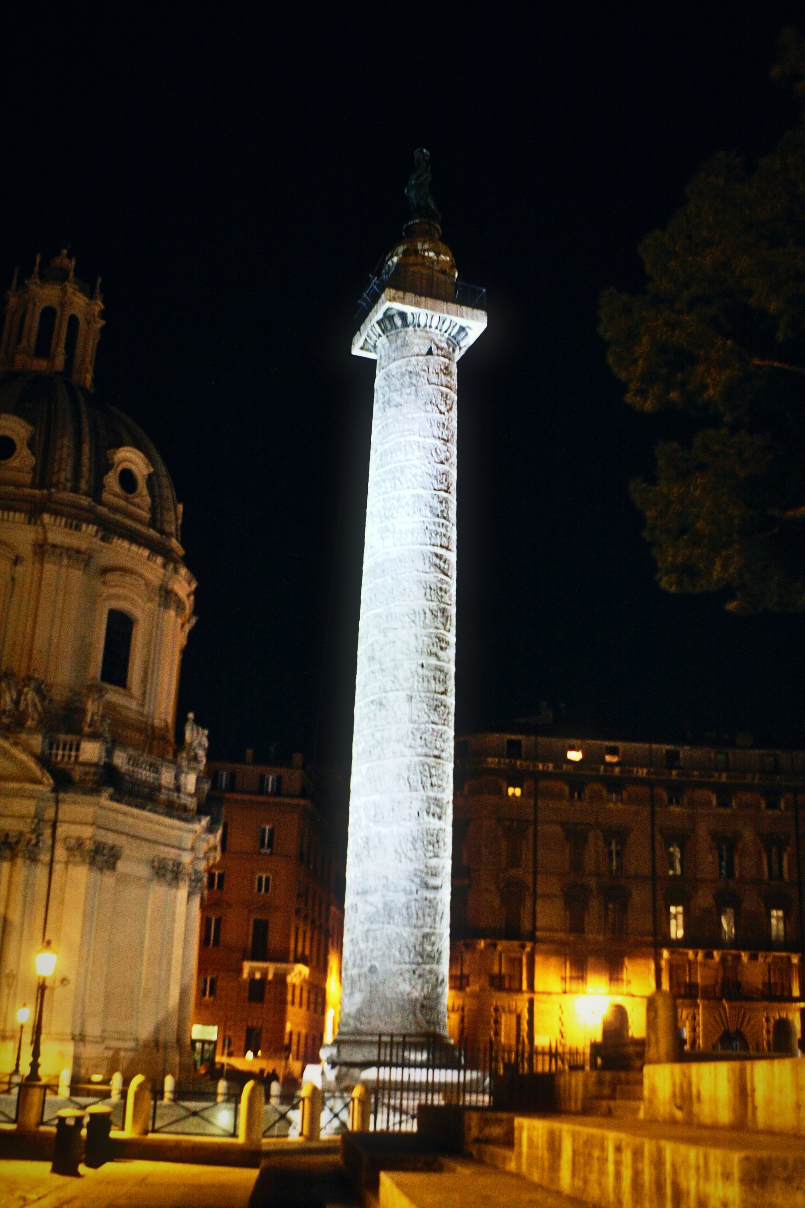 Floodlit carved column rising from a night square beside a domed church.