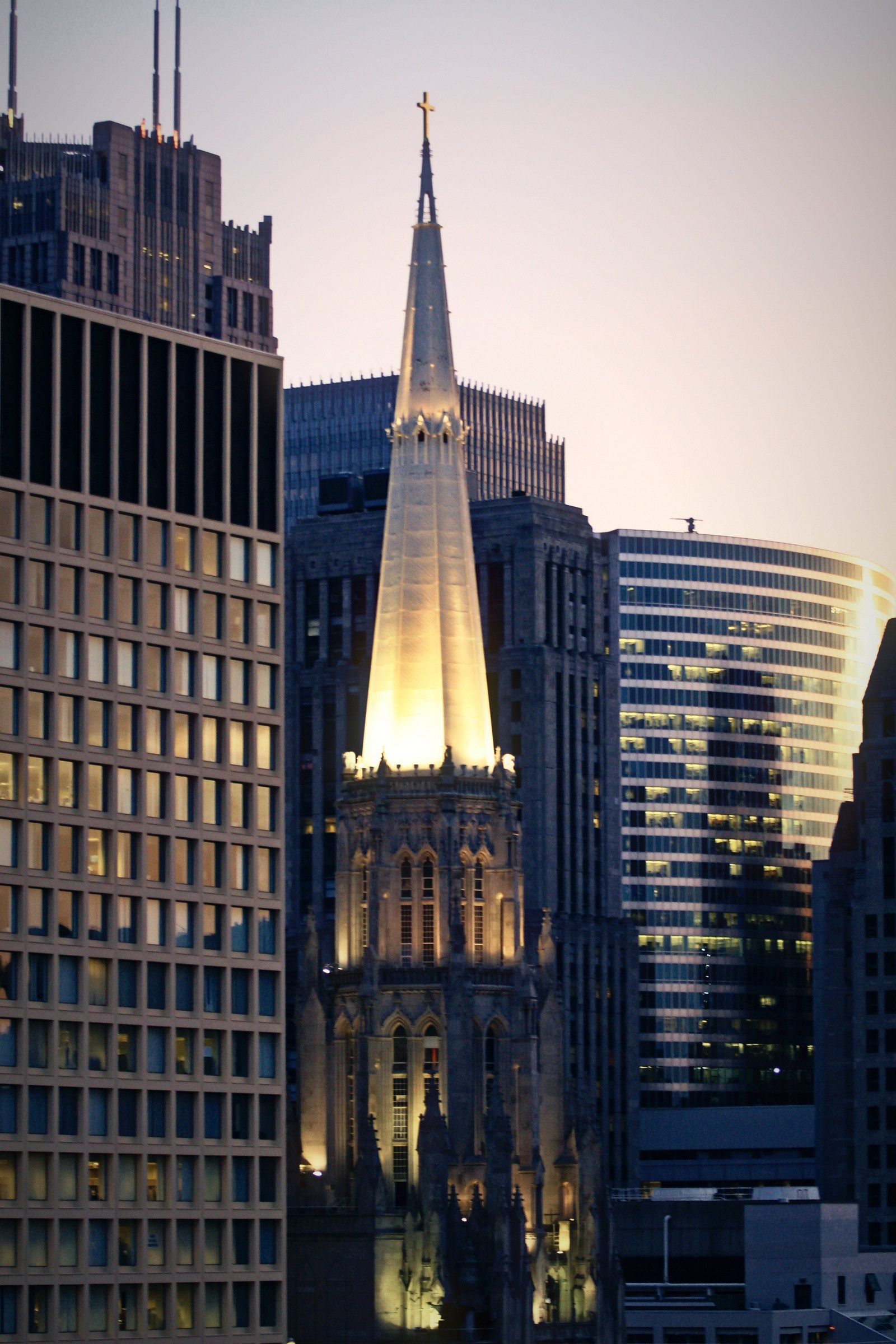 Lit Gothic church spire between office towers under a lilac dusk sky.
