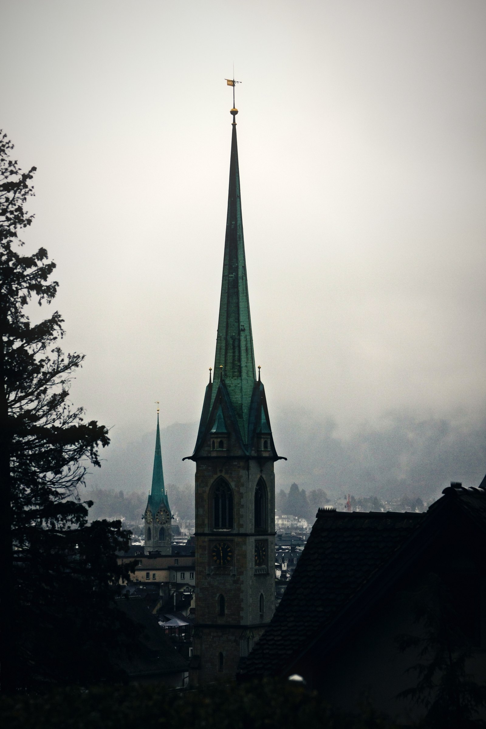 Green church spire rising through mist above dark rooftops and trees.