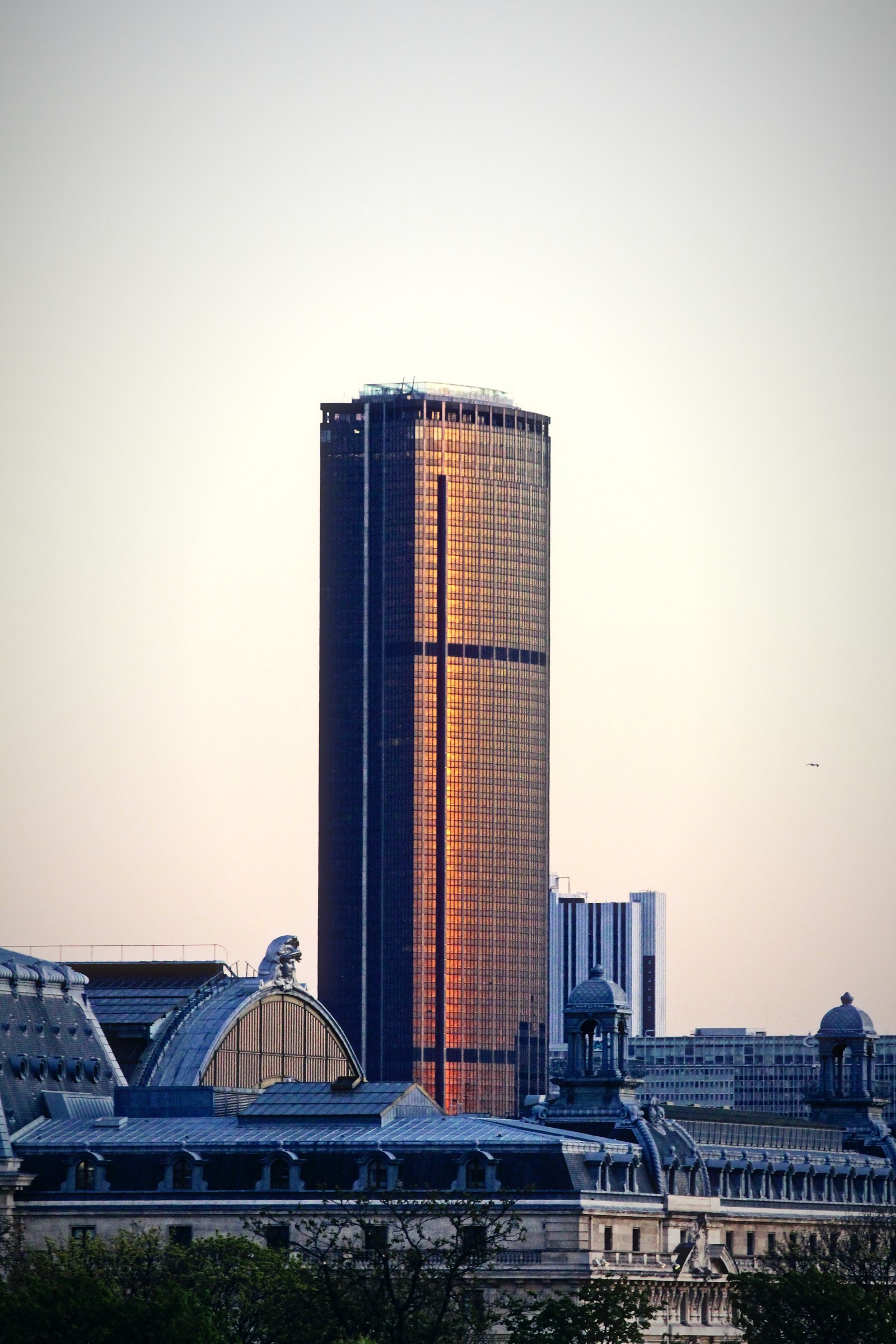 Lone dark skyscraper catching copper evening light above a city of old rooftops.