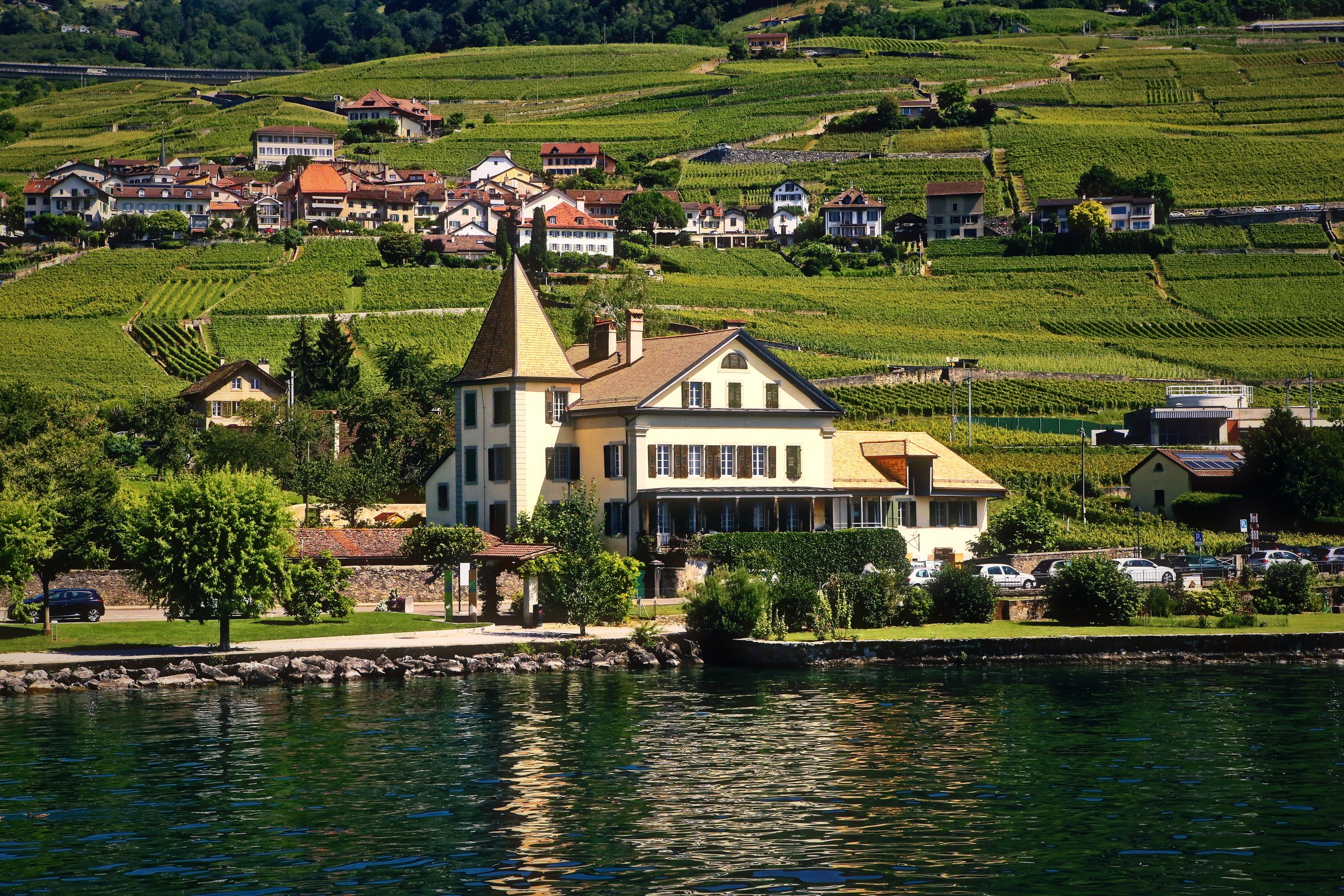 A house with a pointed yellow tower on the lakeshore below terraced vineyards.