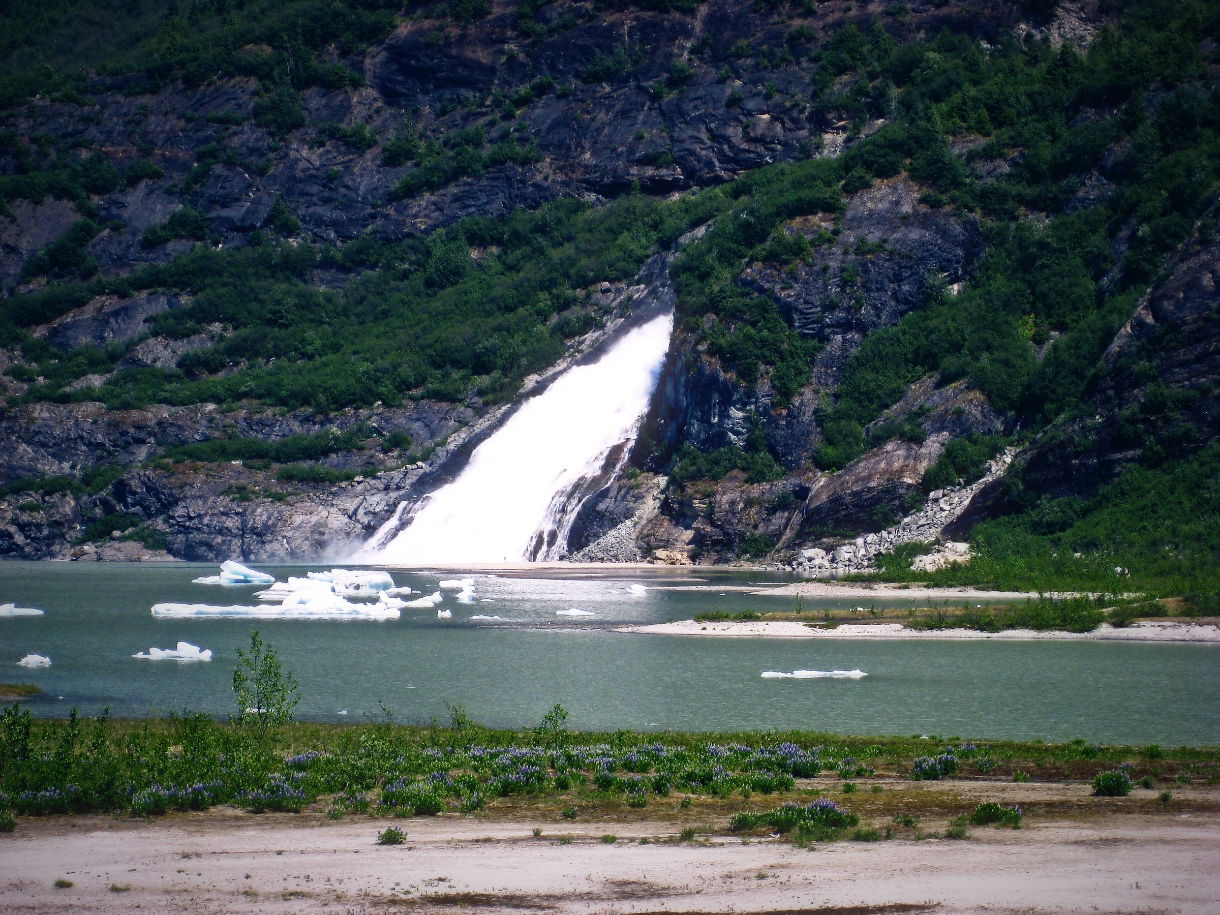 A white waterfall sliding down a dark mountainside into a pale glacial lake.