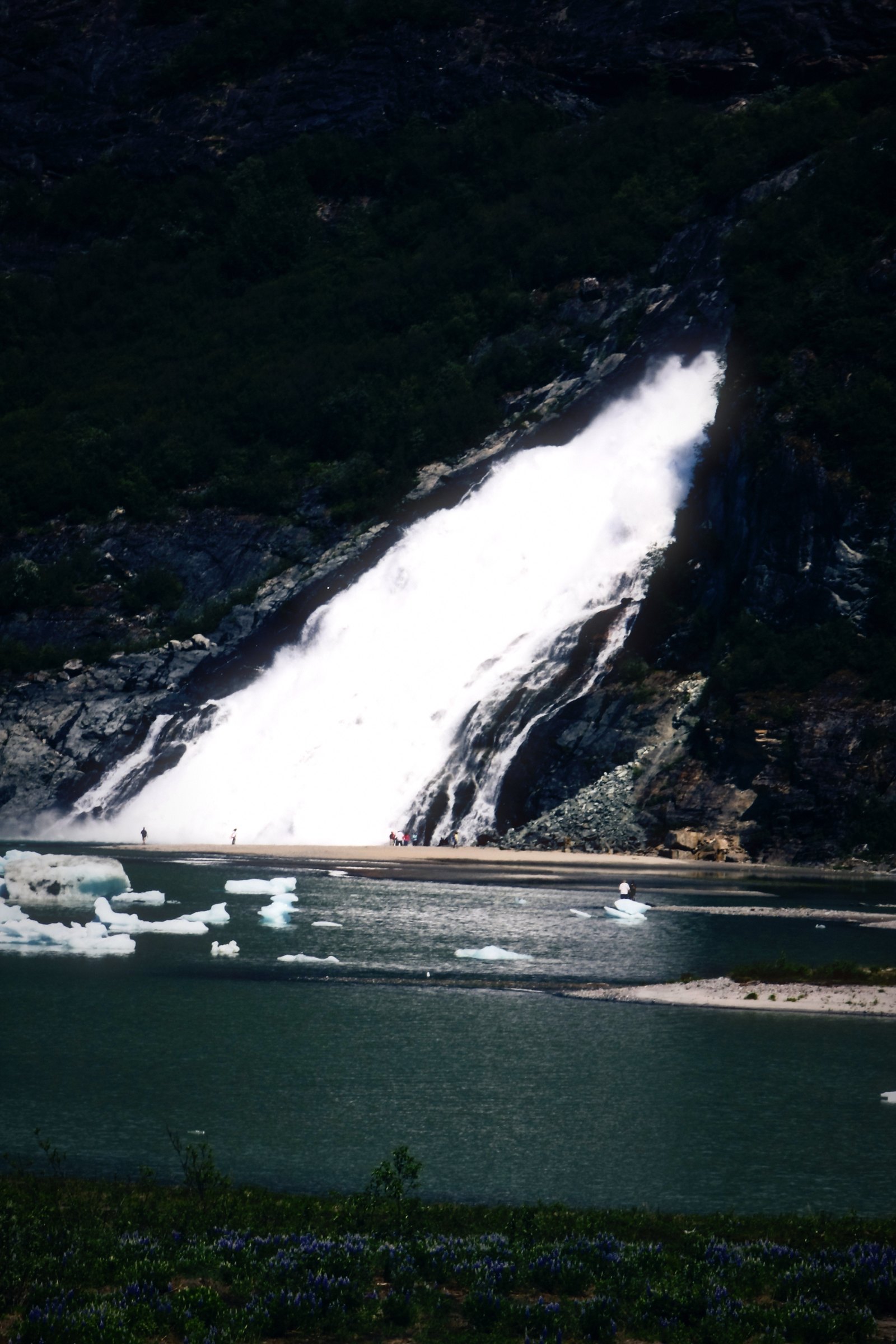 A wide waterfall slanting down a dark rock face into still water.