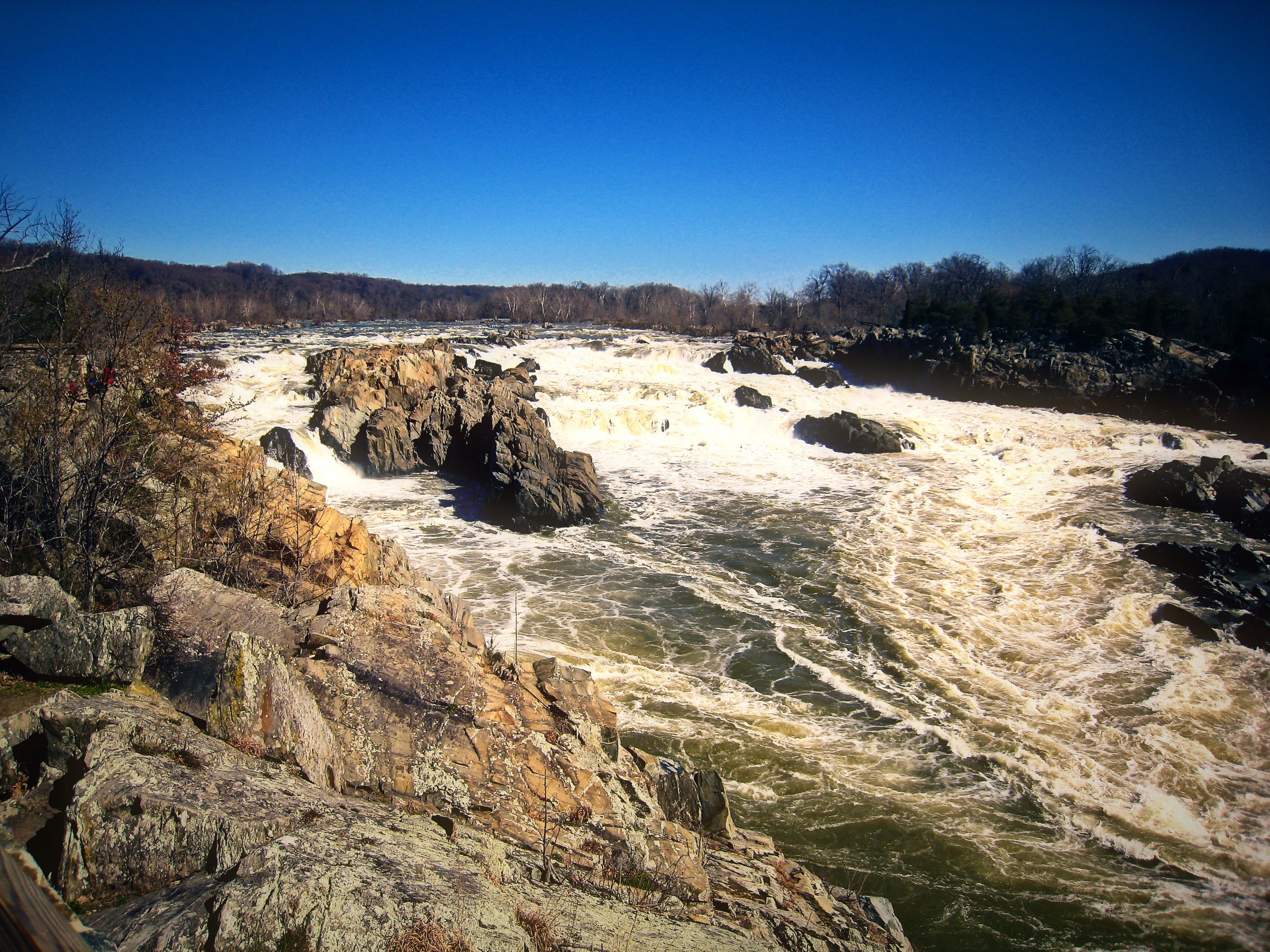 Churning white water spilling between jagged rocks under a clear blue sky.