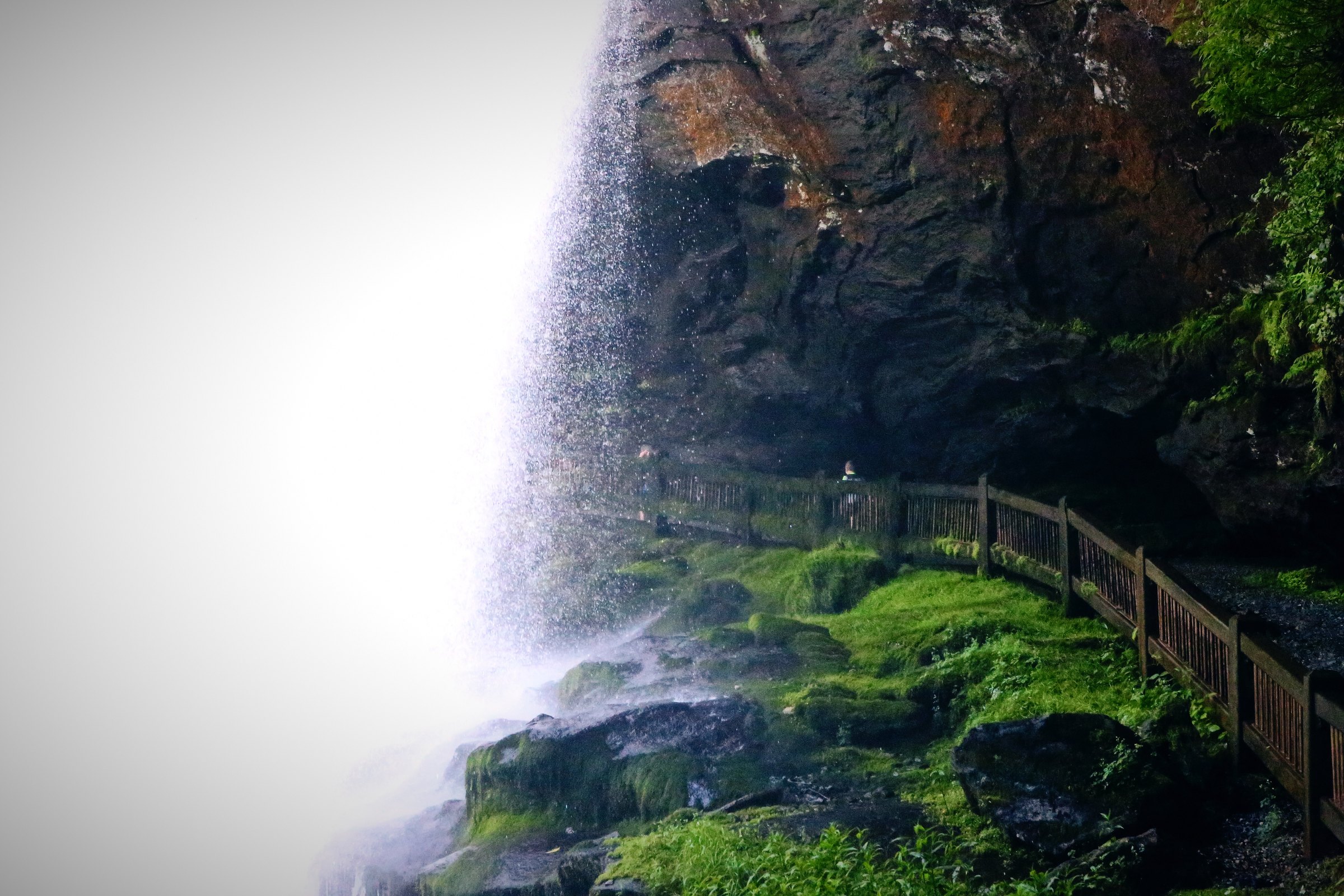 A tall waterfall falling beside a wooden walkway in a green forest gorge.