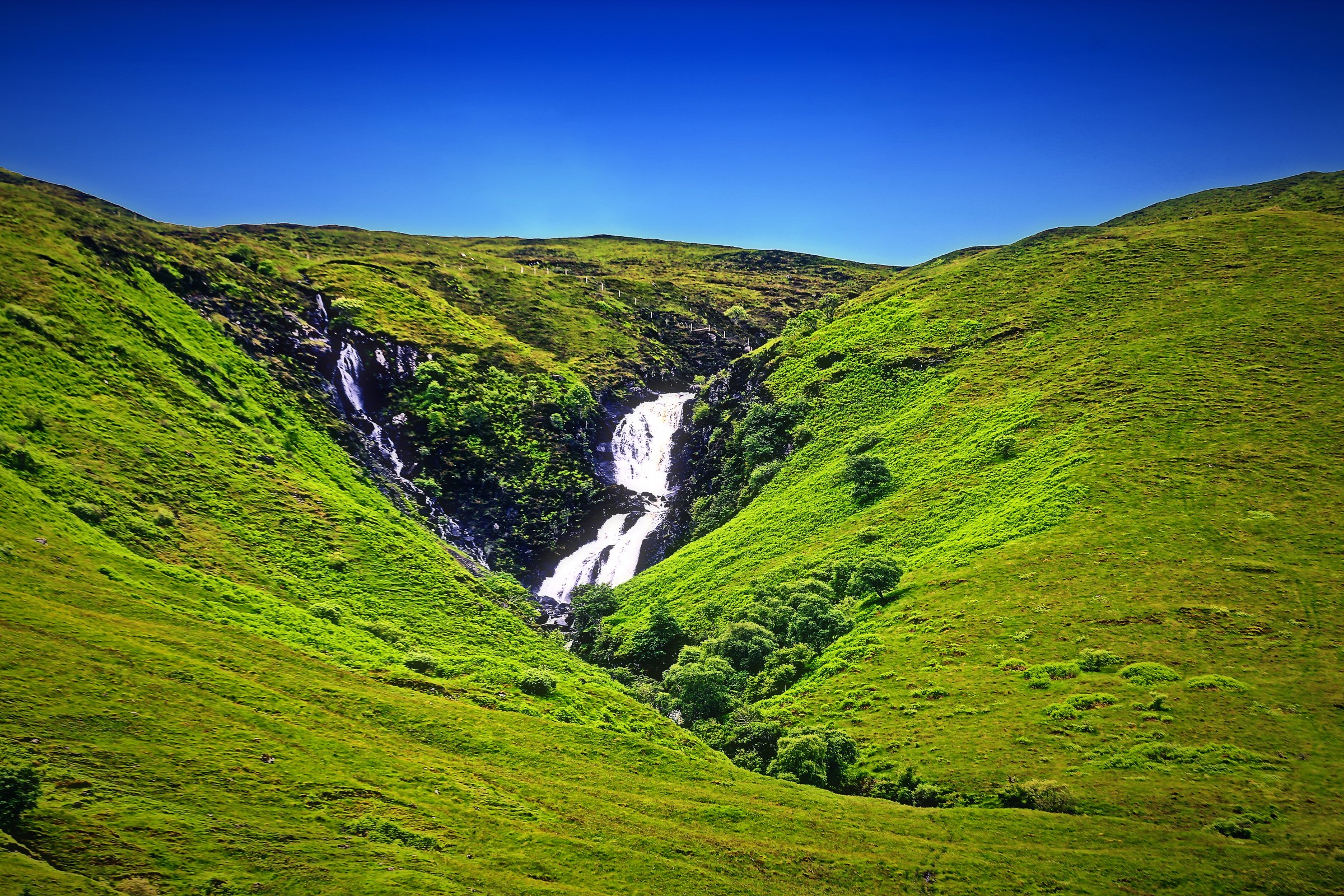 A waterfall dropping down a bright green glen under a deep blue sky.
