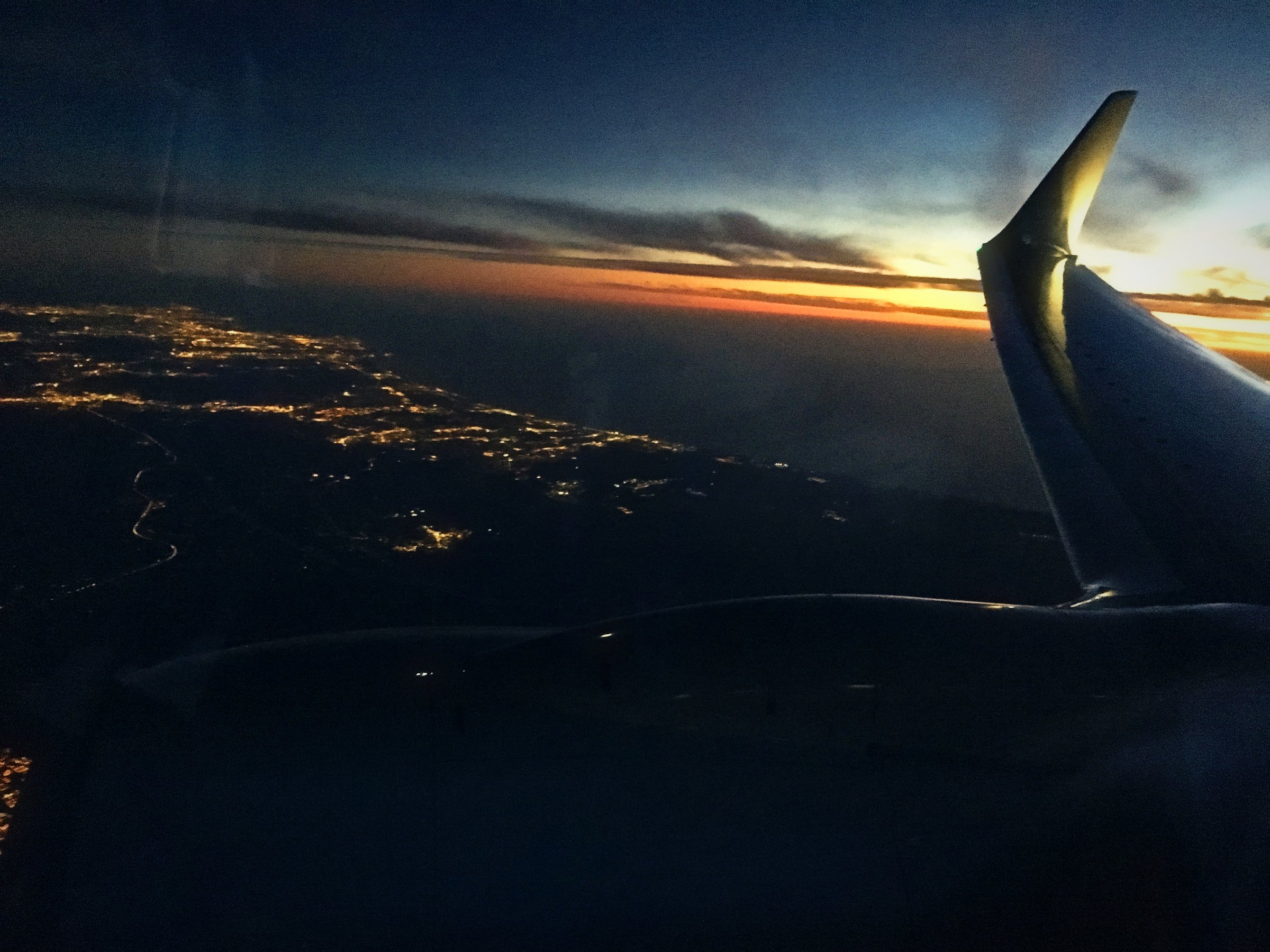 Coastal city lights below an airplane wing as the last orange sunset fades.