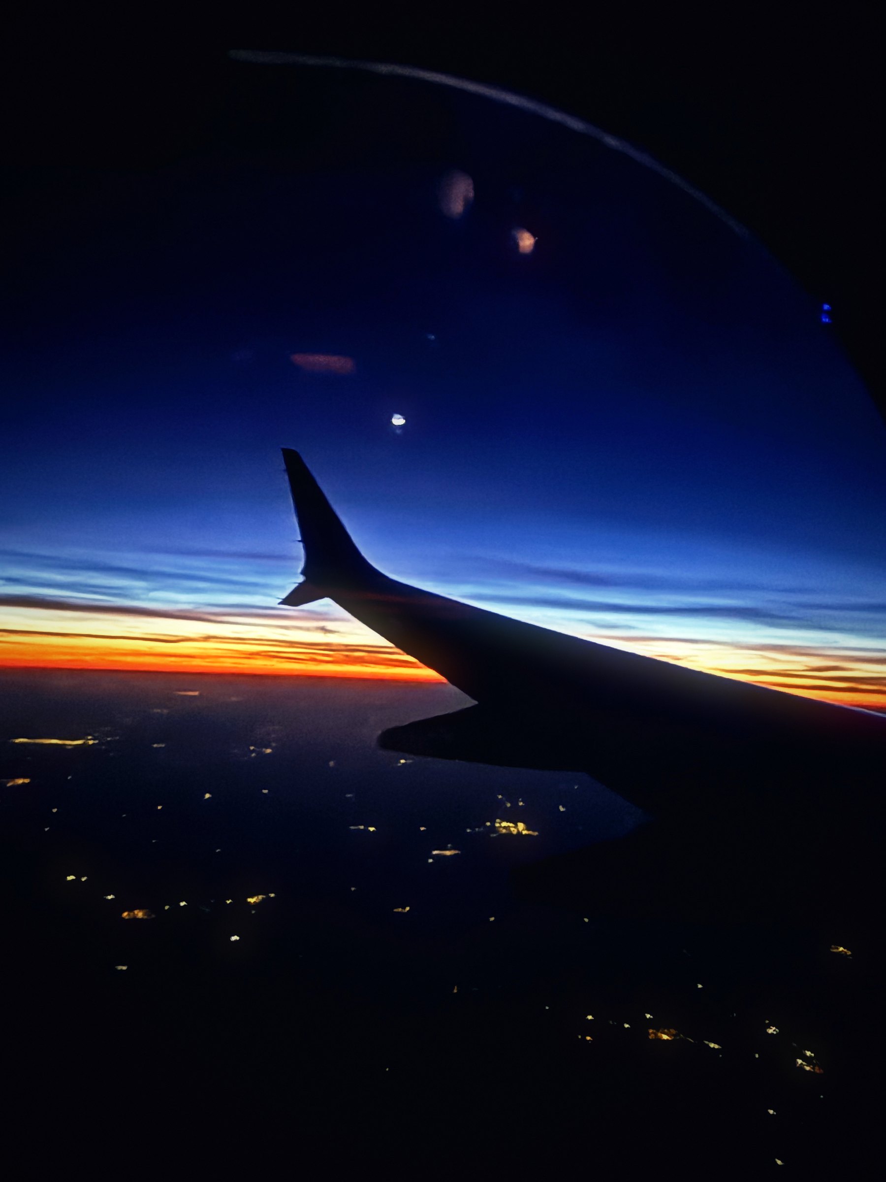 Silhouetted wing at dusk above scattered town lights, with a thin band of sunset on the horizon.