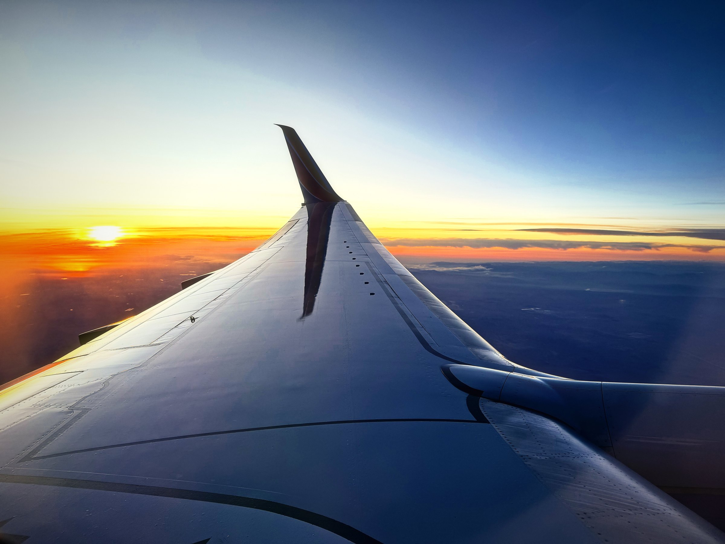 Airplane wing turning silver as the sun sets into a band of orange over the desert.