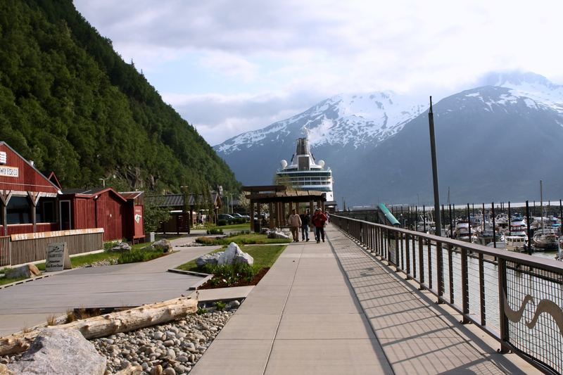Wooden boardwalk with red harbor buildings and snowy peaks beyond.