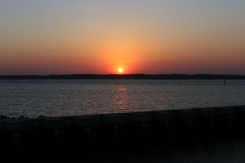 Orange sunset orb over water with twin contrails above.