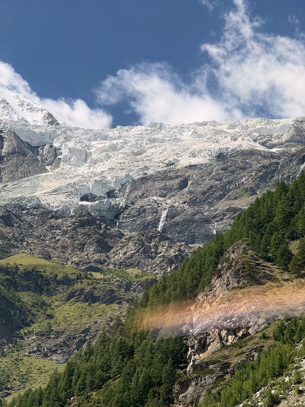 Steep pine-and-snow mountain face with a rainbow flare of light.