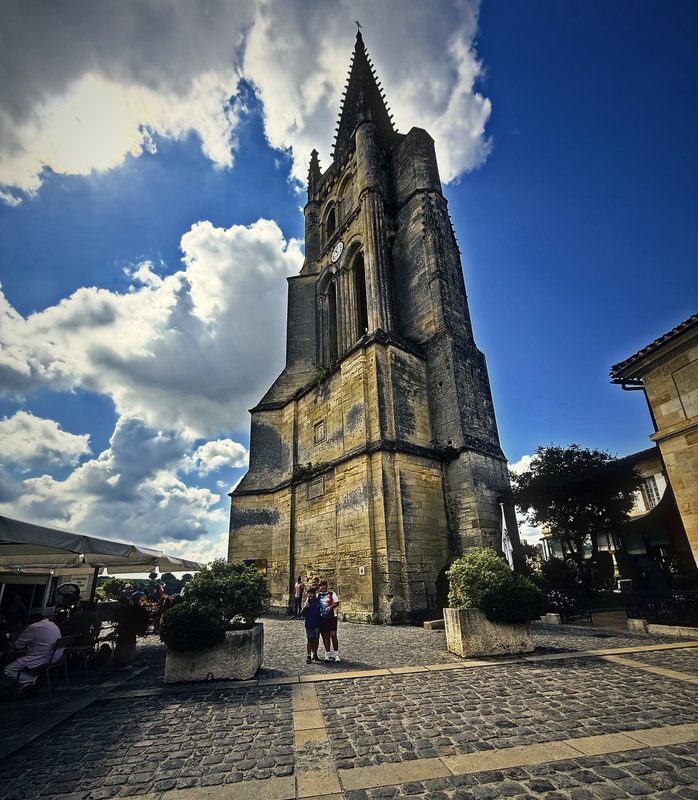 The tall stone bell tower of the Monolithic Church rising over a cobbled square and swirling white clouds.