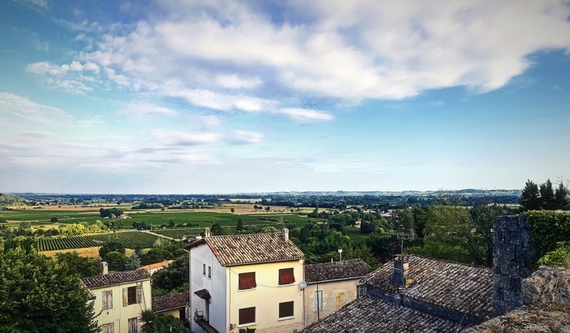 A view over old tiled roofs and a white house toward a wide plain of farmland under bright clouds.
