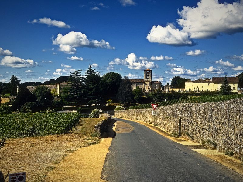 A country road running beside a long limestone wall toward a distant village and tower under scattered clouds.