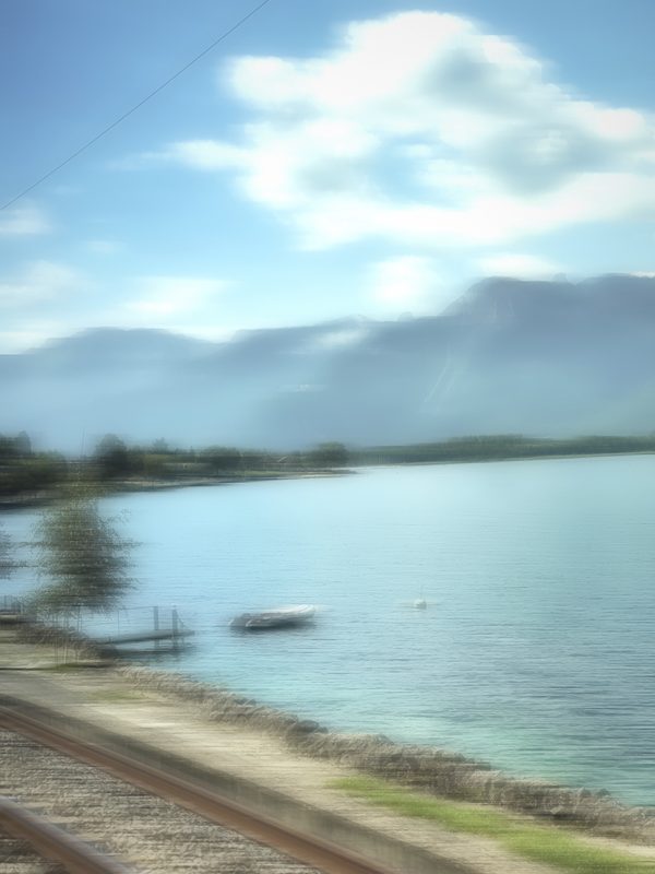 Soft blue lake and mountains blurred past a train window, with a boat on the water.