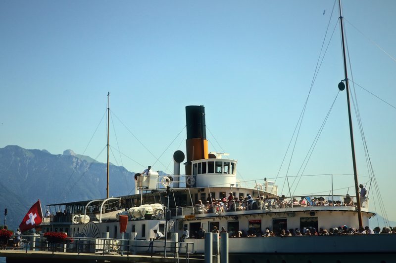 Paddle steamer with a black smokestack and tall mast against the Alps across the lake.