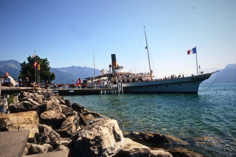 Paddle steamer at its berth framed by shoreline rocks, with hazy mountains behind.