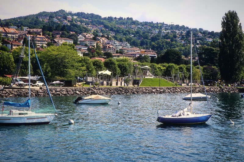 Small sailboats moored on calm water below hillside houses and vineyards.
