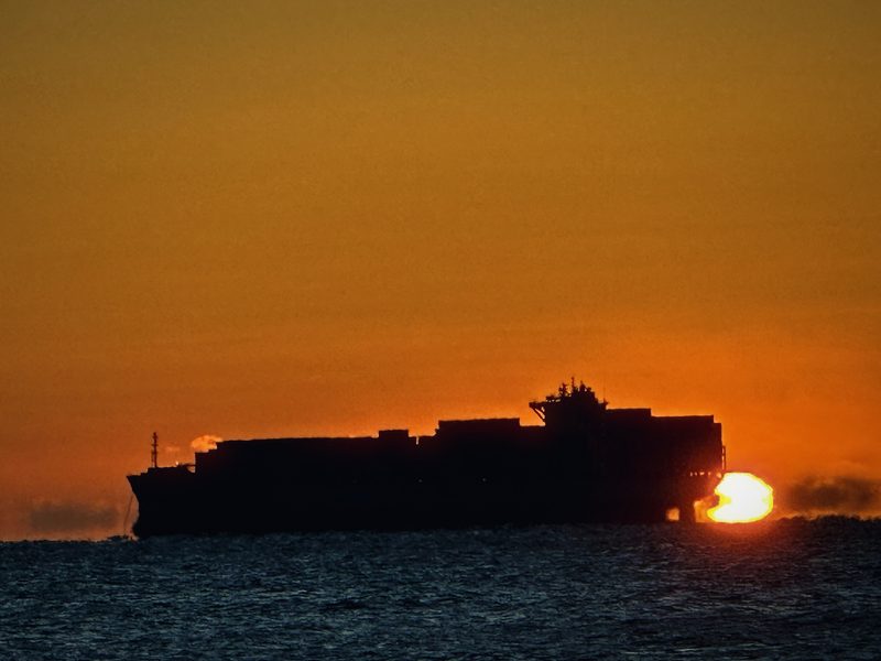 Container ship in silhouette crossing a deep orange sky as the sun touches the sea.