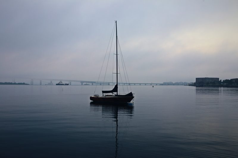 Lone sailboat on glassy, misty grey water with a faint shoreline.