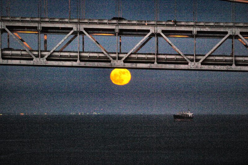 A huge orange full moon rising beneath the dark steel truss of a bridge, a few ships far off on the water.