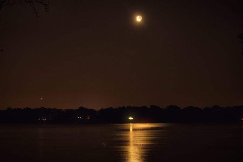 A slim crescent moon with its dark side faintly visible, high above a calm river reflecting a column of light, the sky glowing amber.