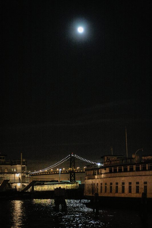 A bright full moon high above San Francisco Bay at night, the Bay Bridge's lights strung low along the water.
