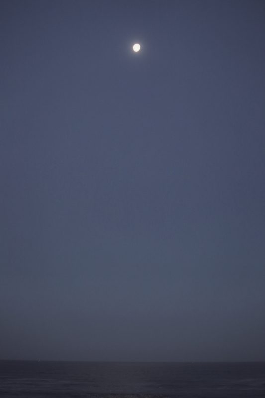 A small moon sinking through soft blue morning sky toward a calm grey sea.