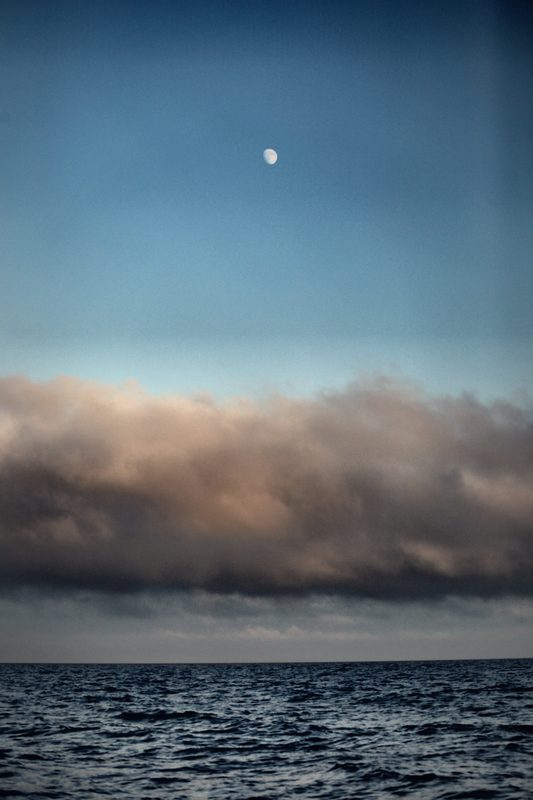 A pale gibbous moon in a clear blue sky above a long bank of fog lying on the sea.