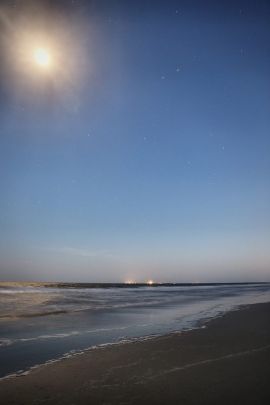 A bright moon high over a wide beach at blue hour, wet sand and calm surf below.