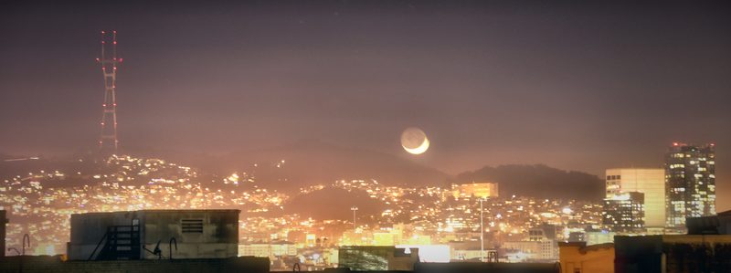 A thin crescent moon with earthshine, hanging over hills of glittering city lights in an amber haze.