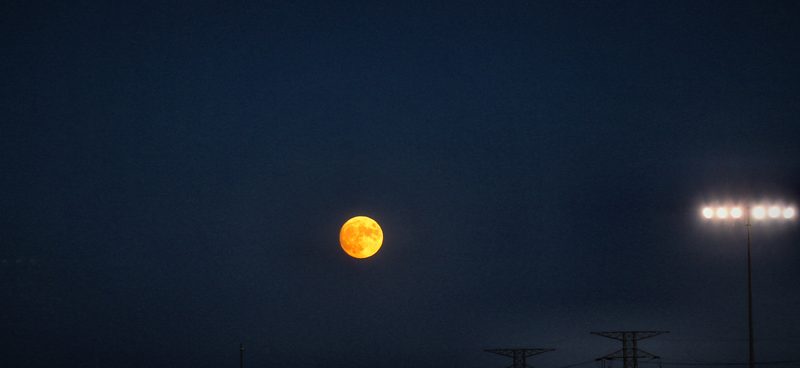 An orange full moon rising in a deep blue dusk beside a bank of field floodlights, the tips of power pylons just below.