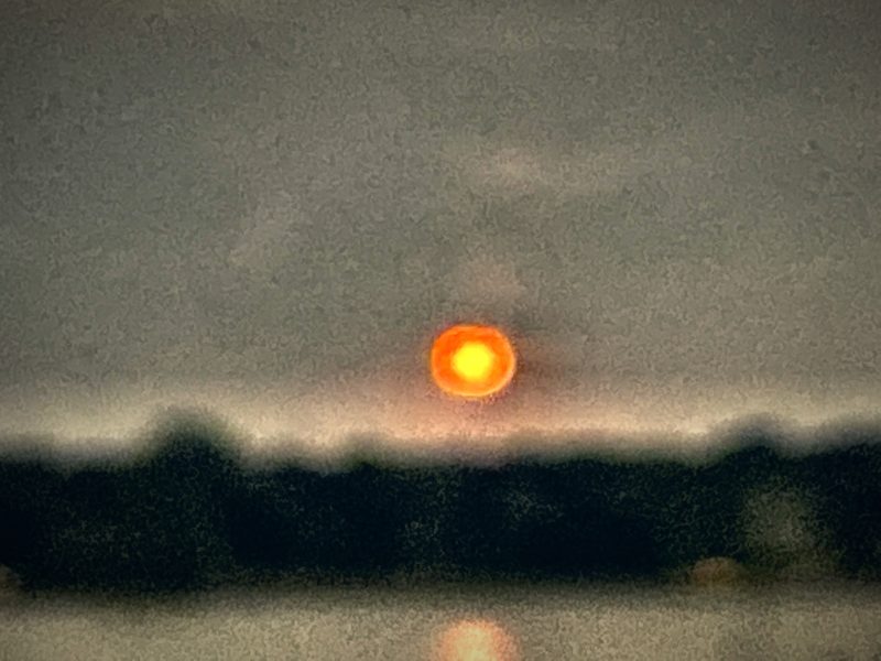 A soft, glowing red-orange moon rising over a dark band of trees beside a lake.